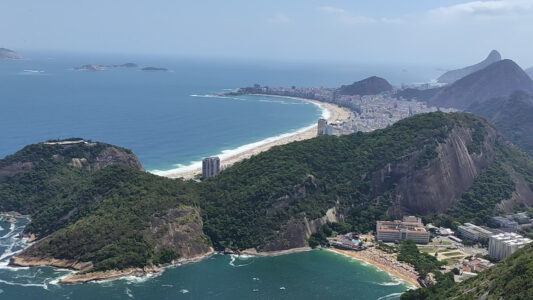 Copacabana e sua curva famosa. Em primeiro plano, a pequena Praia Vermelha, a poucos passos do bondinho do Pão de Açúcar ou da trilha para seu irmão menor, o Morro da Urca. copacabana